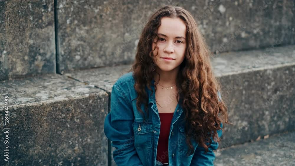 Portrait of the young pretty caucasian teenager girl looking at the side, then turning her face to the camera and smiling while sitting outdoor on the stoned steps. Portrait of the young pretty caucasian teenager girl looking at the side, then turning her face to the camera and smiling while sitting outdoor on the stoned steps.