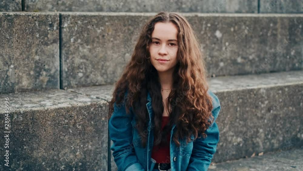 Portrait shot f the cute and stylish teen Caucasian girl with long hair sitting on the stoned steps outside, looking at the side and turning her face to the camera with a smile. Portrait shot f the cute and stylish teen Caucasian girl with long hair sitting on the stoned steps outside, looking at the side and turning her face to the camera with a smile.