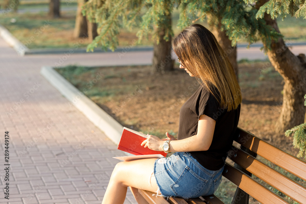 Fototapeta premium A young girl sits on a bench in a park and makes notes. Dressed in a free style. Business woman talking on the phone with customers. 