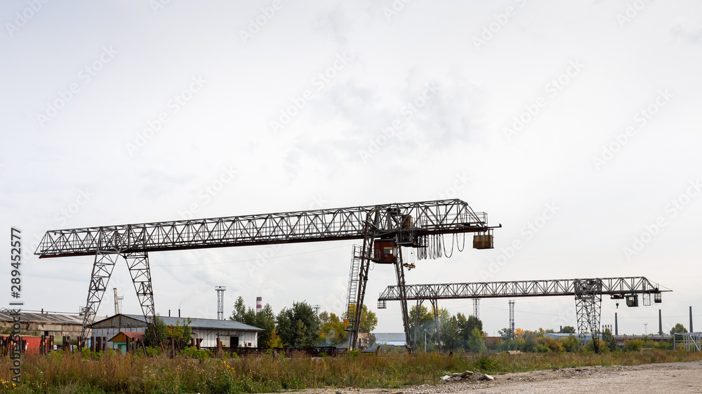 Two large metal gantry cranes at a construction site, in the background industrial warehouses for storing goods. Type of bearing metal structures of gantry crane against the blue sky Two large metal gantry cranes at a construction site, in the background industrial warehouses for storing goods. Type of bearing metal structures of gantry crane against the blue sky