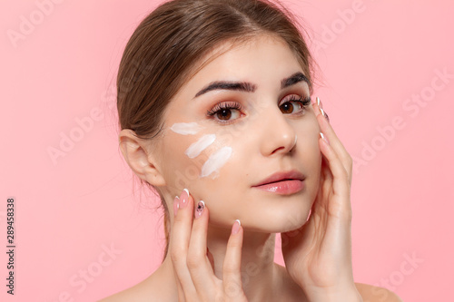 Beautiful model applyes moisturizing cosmetic product to skin holding white tube of a hyaluronic cream isolated over pink background. Concept of beauty and health treatment.