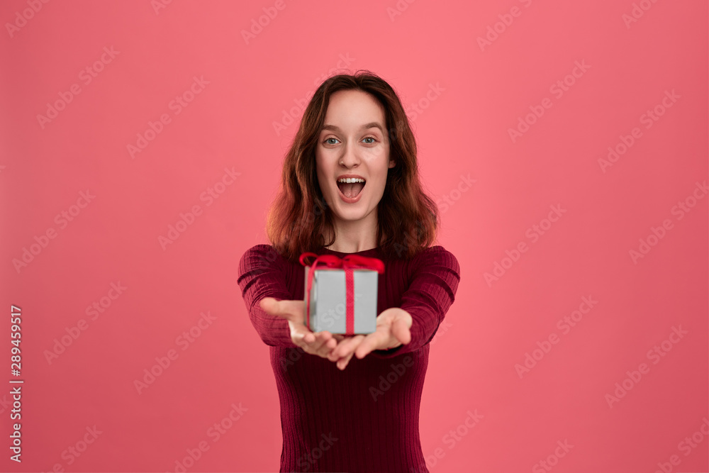 Excited pretty brunette girl holding present box with a ribbon with two hands standing isolated on a dark pink background and smiling at the camera. Celebrating special event. Excited pretty brunette girl holding present box with a ribbon with two hands standing isolated on a dark pink background and smiling at the camera. Celebrating special event.