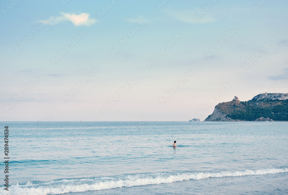 Fototapeta premium a boy into the water looking yacht and calm seascape view of ( sella del diavolo ) in the City of Cagliari from Poetto beach - Sardinia - Italy.