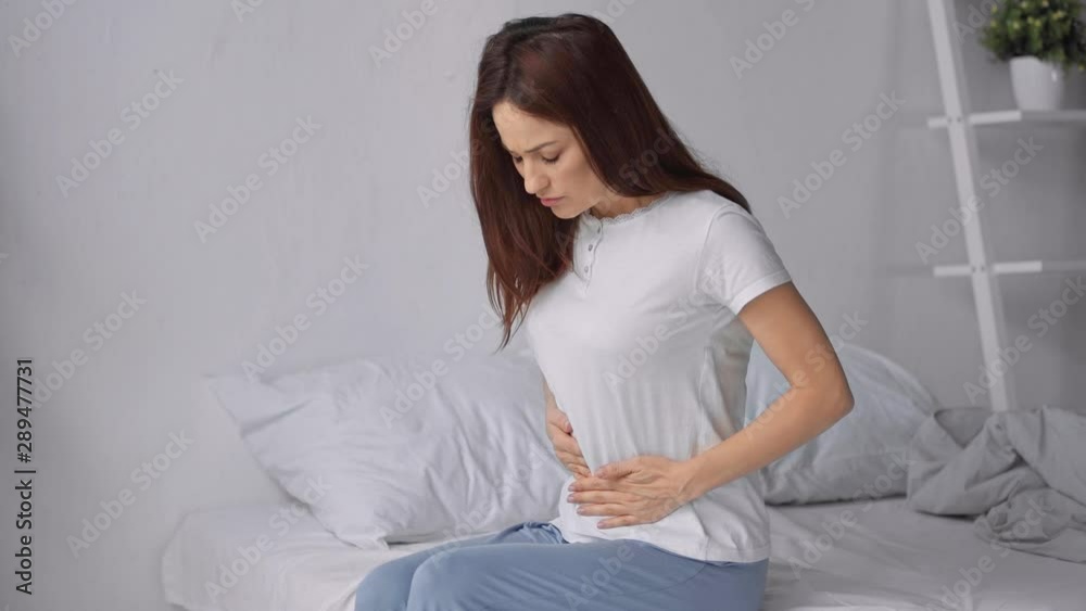 woman sitting on bed in pajamas with stomach ache woman sitting on bed in pajamas with stomach ache