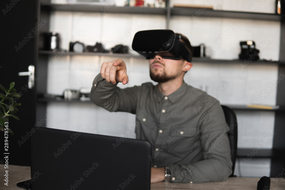 Young bearded man sitting against computer with virtual reality glasses trying to touch something or tap some thing in front of what he seeing. Modern technologies. The concept of future technology. Young bearded man sitting against computer with virtual reality glasses trying to touch something or tap some thing in front of what he seeing. Modern technologies. The concept of future technology.