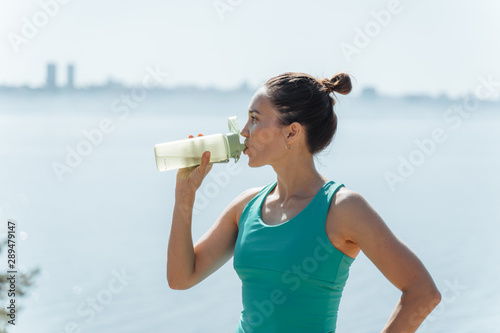 athletic woman drinks water from a bottle on a river background.