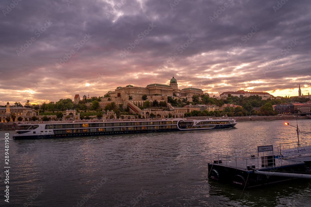 Buda castle with a river cruise passing by on the Danube with dramatic sunset sky