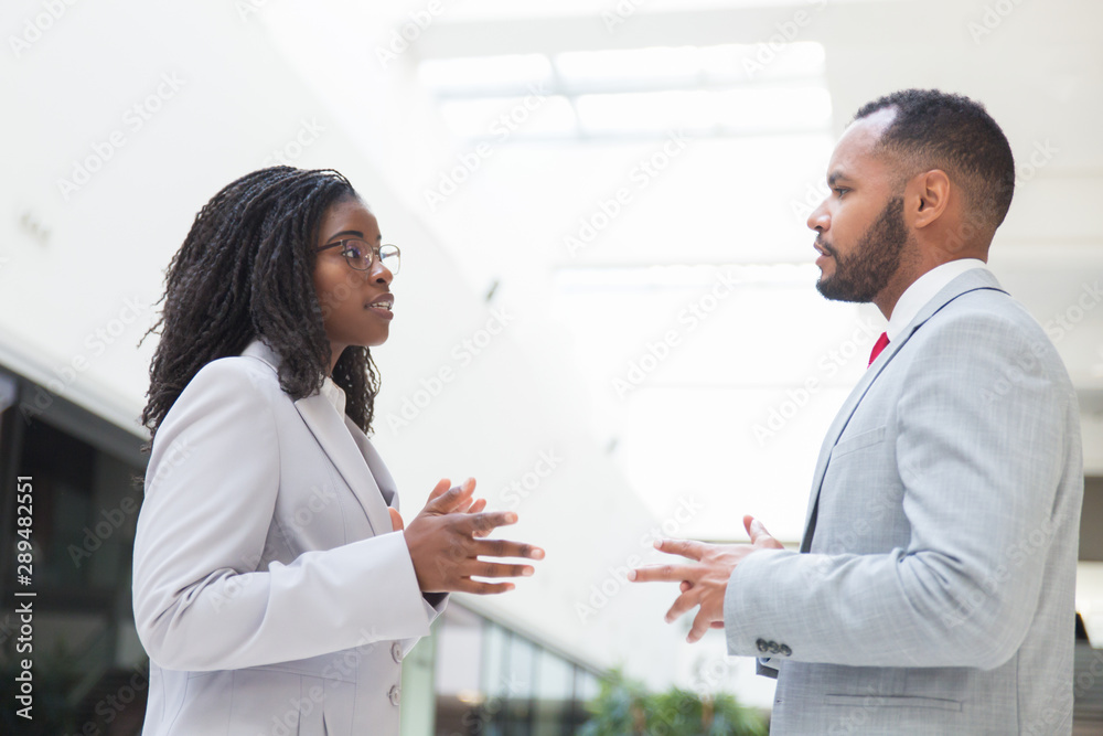 Serious excited business partners arguing about deal in office hall. Business man and woman standing in hallway, talking and gesturing. Negotiation concept