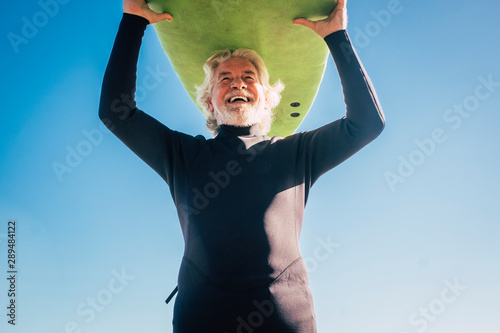 happy senior with surftable on his head is smiling and laughing - old and mature man having fun surfing with a black wetsuits - active retired adult doing activity alone