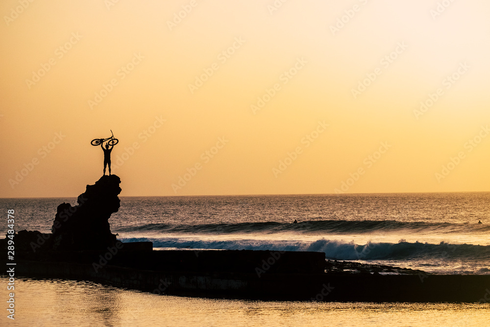adult holding on his arms up a bike at the beach on a big rock - freedom concept and lifestyle - great sunset - ocean and sea with waves with surfers at the background