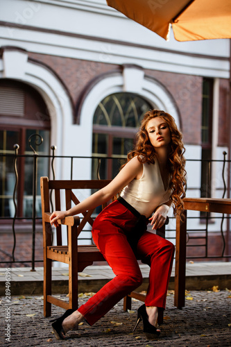 Young curled girl weared in classic trouser jumpsuit on background of old City. Fashion style girl with long curled red hair. Autumn or early spring in the City. Street style Modern strict urban girl.