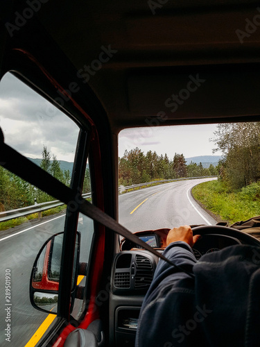 Image from passenger compartment of car driving along road among green trees in Norway on summer.
