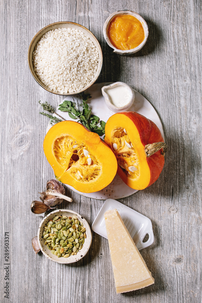 Ingredients for cooking pumpkin risotto. Raw uncooked risotto rice in ceramic bowl, sliced pumpkin, cream, seeds, parmesan cheese, garlic and herbs over grey wooden background. Flat lay, space