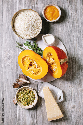 Ingredients for cooking pumpkin risotto. Raw uncooked risotto rice in ceramic bowl, sliced pumpkin, cream, seeds, parmesan cheese, garlic and herbs over grey wooden background. Flat lay, space