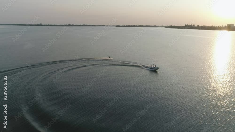 view from above, man in life vest rides on wakeboard along river holding rope tied to motorboat on background of sunset and port in slow motion outdoor