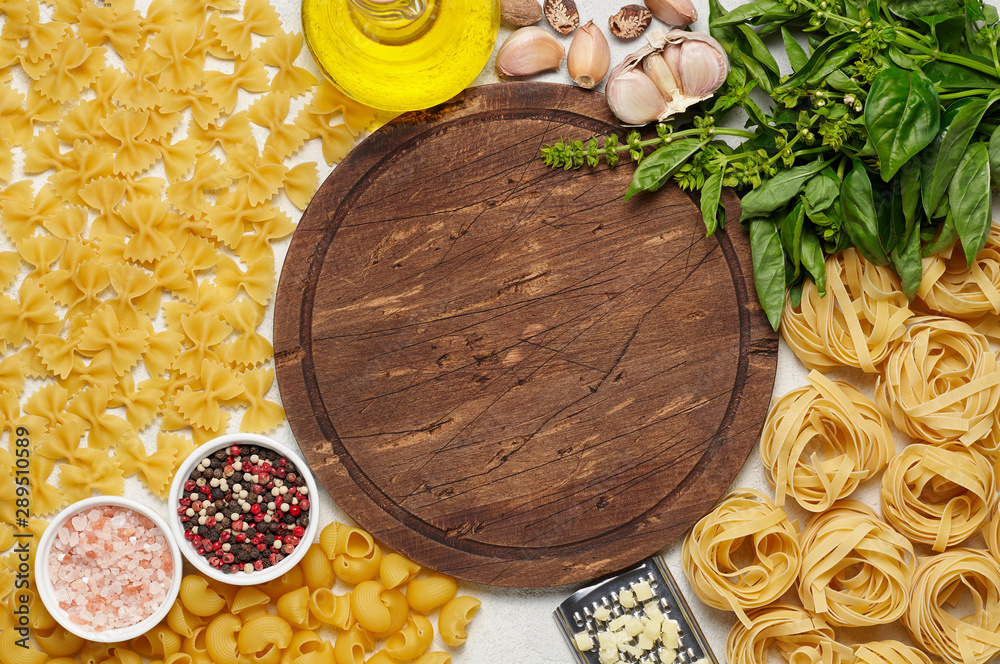 Round rustic wooden cutting board surrounded by different types of Italian pasta