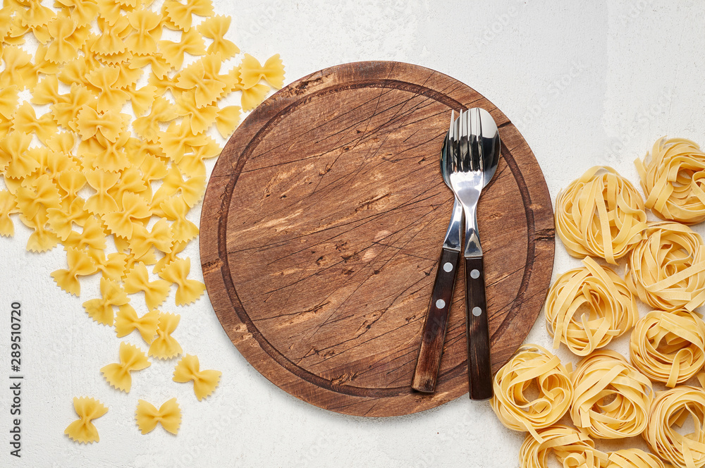 Fork and spoon on round rustic wooden cutting board surrounded by different types of Italian pasta