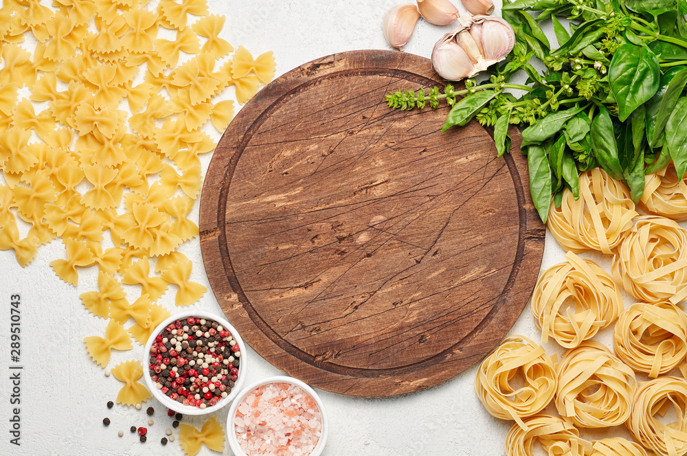 Round rustic wooden cutting board surrounded by different types of Italian pasta
