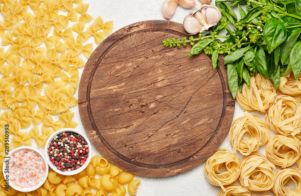 Round rustic wooden cutting board surrounded by different types of Italian pasta