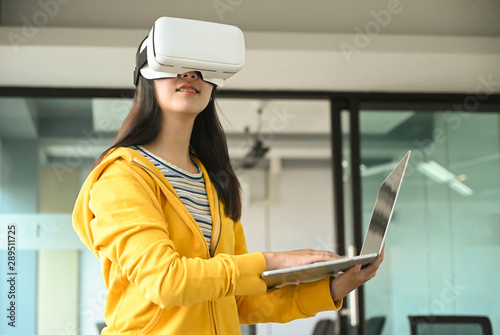 Asian girl wearing a yellow shirt, using VR headset. She is holding laptop and typing a keyboard.