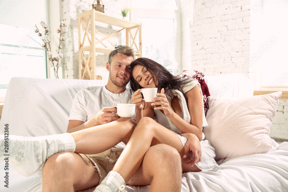 Couple of lovers at home relaxing together. Caucasian man and woman having weekend, looks tender and happy. Concept of relations, family, autumn and winter comfort. Drinking tea and hugging. Couple of lovers at home relaxing together. Caucasian man and woman having weekend, looks tender and happy. Concept of relations, family, autumn and winter comfort. Drinking tea and hugging.