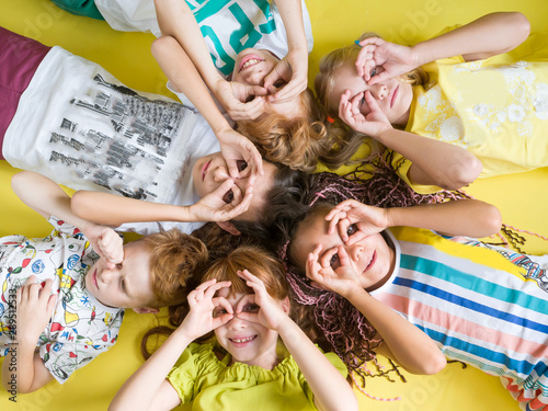 Fashionable joyful children in bright summer clothes with their hands to their eyes lie cheerfully on a yellow background looking up. Happy children
