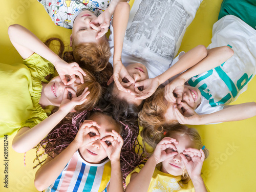 Beautiful joyful children with their hands to their eyes lie cheerfully on a yellow background looking up. Happy children