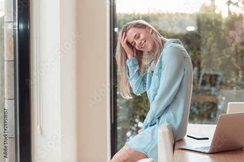 Charming young smiling woman enjoys her vacation in a tropical country while spending her morning in a chic hotel room with a laptop. The concept of luxury relaxation for the millenials