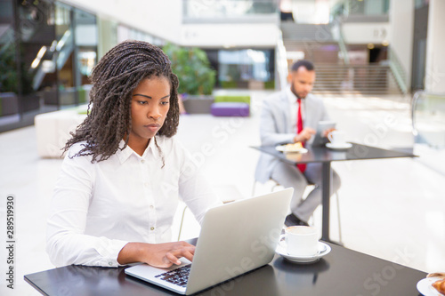 Focused hardworking female professional working on report during coffee break. Young African American business woman using laptop, man using tablet in background. Co-working concept