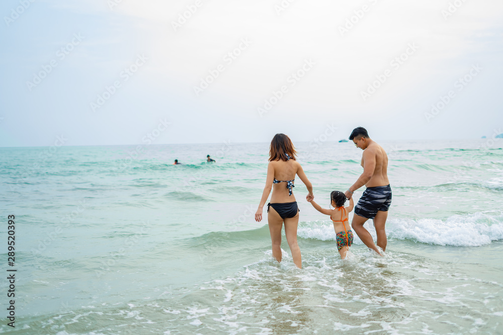 Happy asian family - father, mother, baby daughter hold hands and run with fun along edge of sea on sand beach. Active parents and people outdoor activity on tropical summer vacations with children.