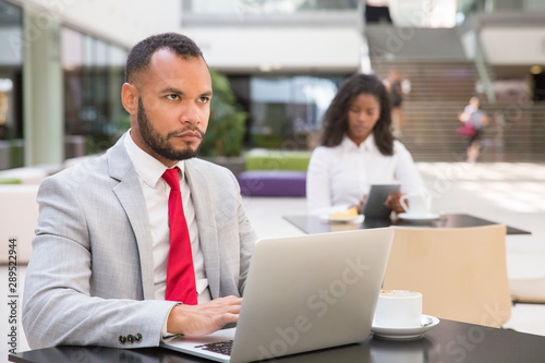 Serious business leader working on laptop in office cafe. Young black woman drinking coffee and using tablet in background. Wireless technology concept