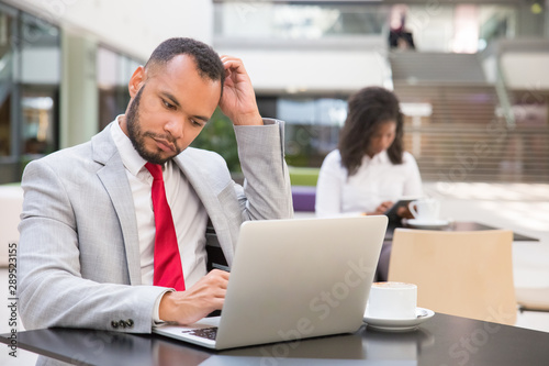 Pensive male manager using laptop while drinking coffee in office lobby. Young black woman using gadget in background. Digital technology concept