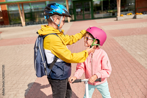 safety, childhood and care concept - happy school boy with backpack fastening girl's helmet for scooter riding