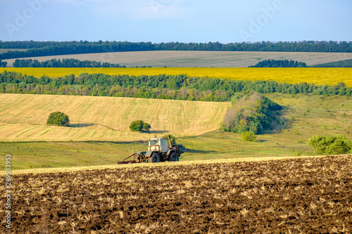 An agricultural tractor plows a field with a plow after harvested wheat. In the background are fields of sunflowers.