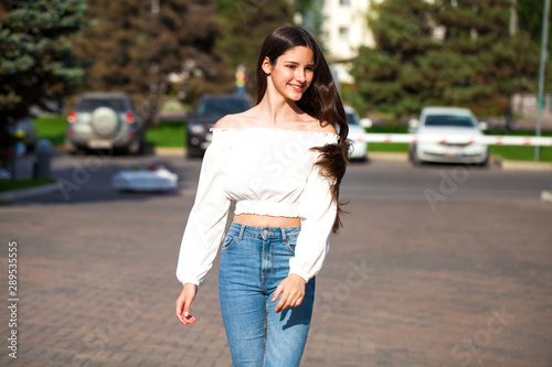 Young beautiful brunette woman in jeans and white blouse walking in summer street