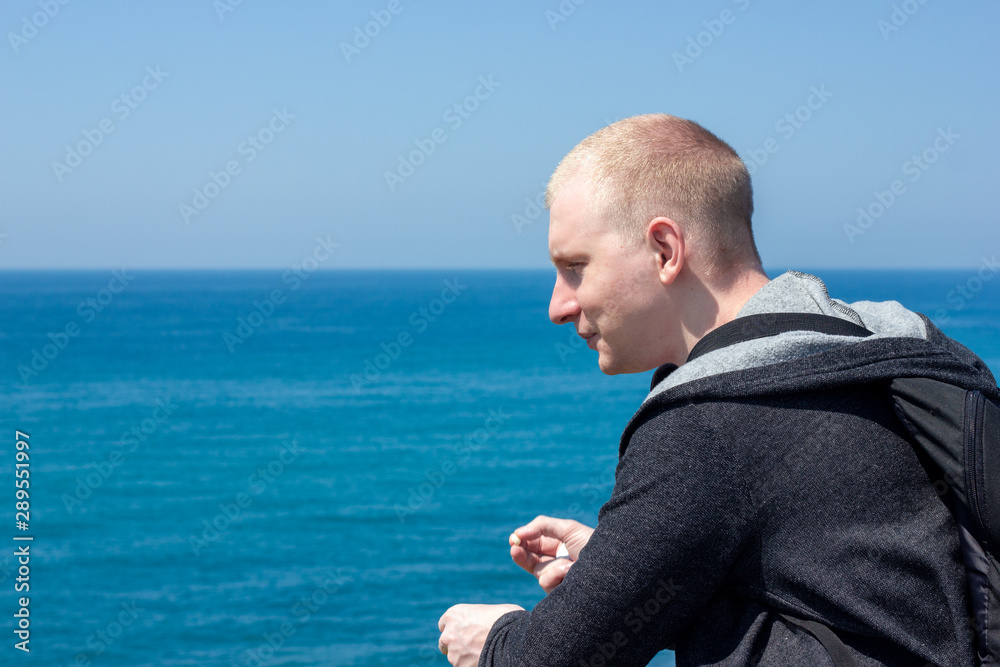 young short-haired man stands on the shore of the blue sea and smiles on a sunny summer day