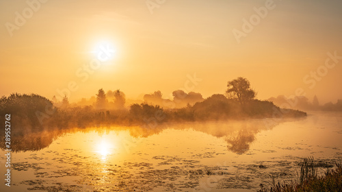 Beautiful panorama of the golden hour with misty river, reflected trees and rising sun. Foggy morning background in golden tones