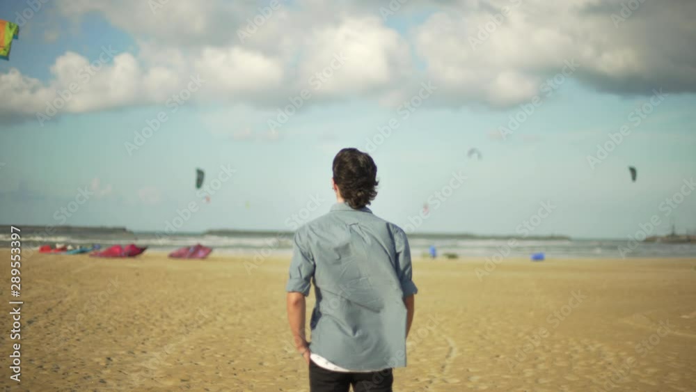 Young sensitive guy looking at kitesurfes flying in the sky under a blue and cloudy sky by the sea. Beach in a windy day. Young sensitive guy looking at kitesurfes flying in the sky under a blue and cloudy sky by the sea. Beach in a windy day.