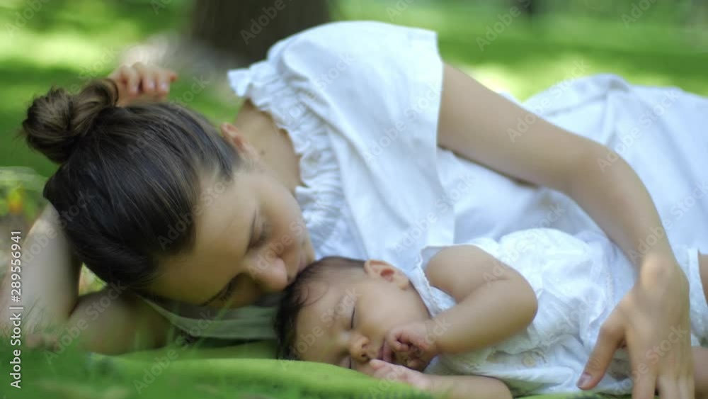 Mother kisses her Baby sleeping on a green grass Outdoors. Happy smiling young Mother and Child in Green Summer Park. Beautiful family in spring park enjoying nature. Mother kisses her Baby sleeping on a green grass Outdoors. Happy smiling young Mother and Child in Green Summer Park. Beautiful family in spring park enjoying nature.