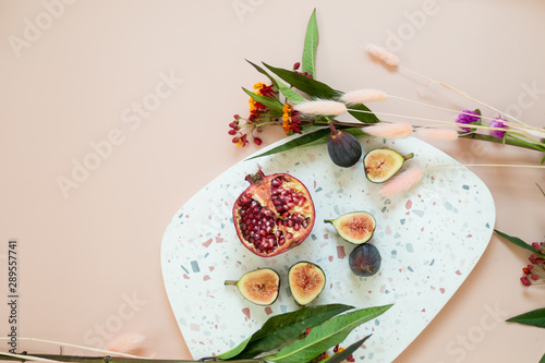 Fresh pomegranate and figs cut open on terrazzo dish, styled with flowers and pink background, copy space