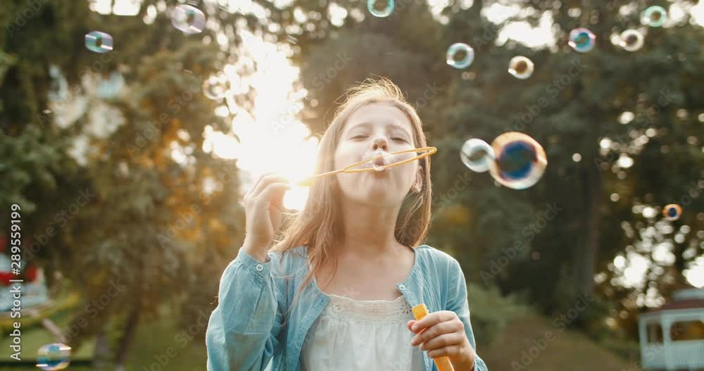 Portrait of the cheerful and smiled beautiful Caucasian girl with long fair hair blowing bubbles in the sunny park among trees. Portrait of the cheerful and smiled beautiful Caucasian girl with long fair hair blowing bubbles in the sunny park among trees.