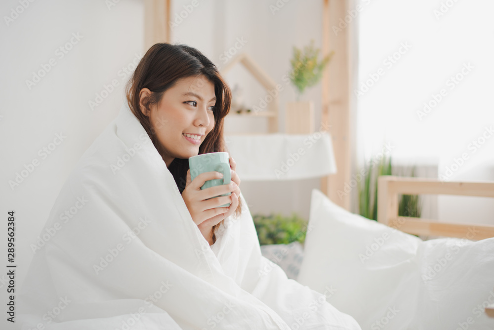 Young beautiful Asian woman sitting on white bed and cover body by white blanket which hole a cup of worm water on hand and smiling and felling happy. flu season concept. Young beautiful Asian woman sitting on white bed and cover body by white blanket which hole a cup of worm water on hand and smiling and felling happy. flu season concept.