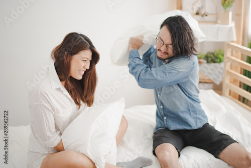 Young couple Asian man and woman sitting on white bed having a fun and felling happy when pillow fighting in bed room at home.