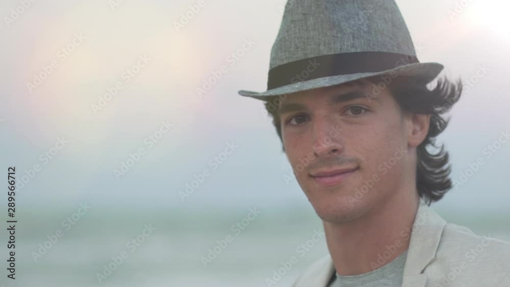 Slow motion portrait of fashionable trendy handsome young man with hat looking at camera and smiling at sunset in the city.