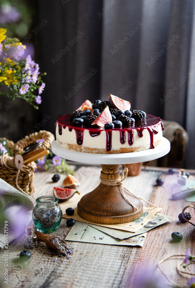 homemade tasty whole cheesecake decorated with figs, blackberries, blueberries and purple sauce on top served on wooden cake stand on grey table with flowers and berries, selective focus