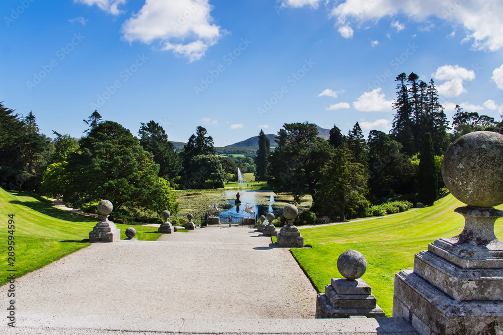 A wide footpath leading to lake with fountain surrounded by green trees, Sugerloaf mountain in the background, Powerscourt gardens, Wicklow, Ireland