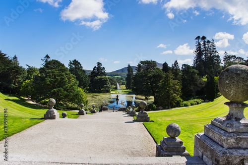 A wide footpath leading to lake with fountain surrounded by green trees, Sugerloaf mountain in the background, Powerscourt gardens, Wicklow, Ireland photo