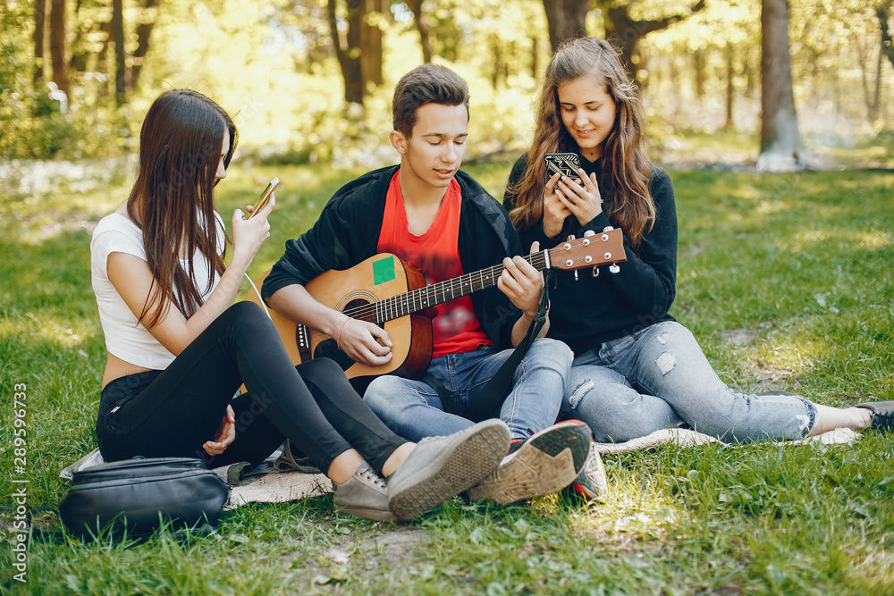 Three friends sitting on a green grass. Boy playing on a guitar with his two girlfriends. Two girls have fun with their friend