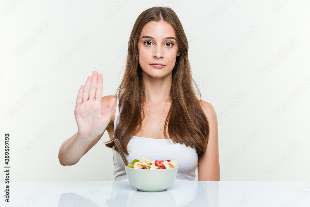 Young caucasian woman eating fruit bowl standing with outstretched hand showing stop sign, preventing you. Young caucasian woman eating fruit bowl standing with outstretched hand showing stop sign, preventing you.