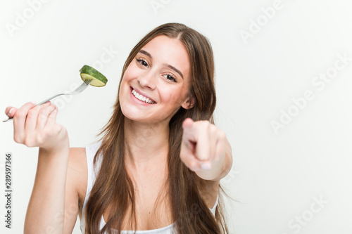 Young caucasian woman eating cucumber cheerful smiles pointing to front.